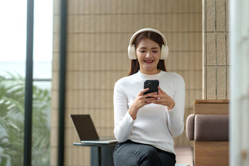 Young woman wearing headphones, smiling and relaxing while using a smartphone for entertainment in a modern indoor space