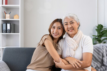 Asian grandmother and adult daughter embracing, sitting on a sofa and smiling, representing love, family bonding, and multigenerational support
