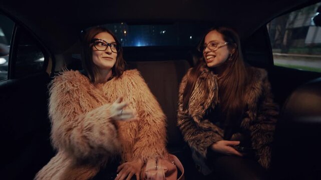 Two stylish girls in luxurious fur coats sit in the back seat of a taxi, laughing and chatting as the bright city lights flash through the windows at night.