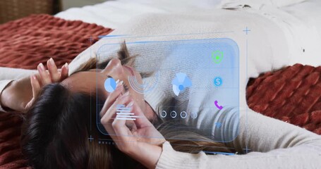 mid-adult woman lying on bed raising smartphone and studying AR chart icons for tech data analysis - Powered by Adobe