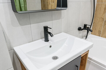 Modern bathroom sink with a black faucet, white basin, and gray cabinet. Tiled walls and a bathtub are visible