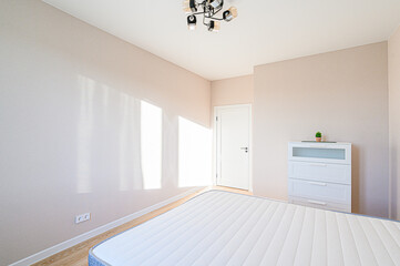 A bright, neutral bedroom with a bed, white dresser, and door. Sunlight streams through a window. Modern ceiling light
