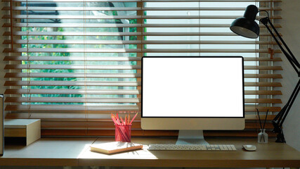 Real photo of bright room interior with wooden desk with lamp, notebooks and empty screen computer standing by the window