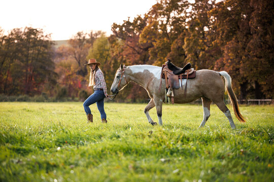 Cowgirl in western clothing leading horse across pasture during autumn sunset. Woman walking with horse, symbolizing slow living and country lifestyle