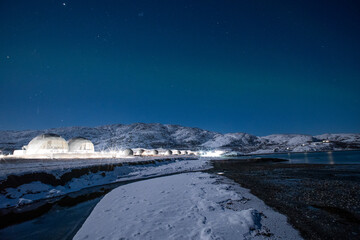 Low-angle view of illuminated geodesic glamping domes on a snowy riverbank, set beneath the Aurora Borealis and star-filled night sky
