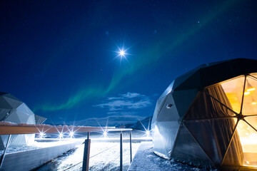 Close-up of two geodesic glamping domes on a snowy deck, with a vibrant band of green Aurora Borealis stretching across the starry night sky