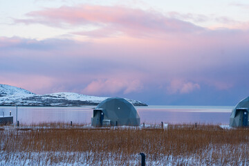 Close-up of a gray geodesic glamping dome with foreground dry grass and a pink-hued twilight sky reflecting over the calm Arctic sea