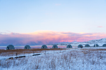 Horizontal row of geodesic glamping domes on a snowy coastal plain under a spectacular arctic sky dominated by pink and purple clouds