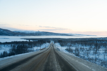 A long, snow-dusted road extends into a vast winter landscape with a forested valley partially obscured by a layer of fog