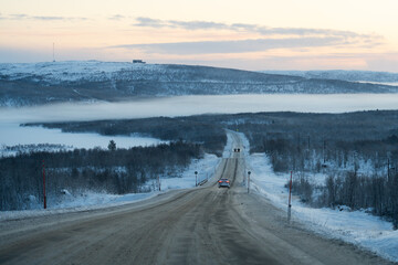 A white car drives down a cleared, snow-covered road toward a forested valley partially hidden by a thick layer of fog and low clouds