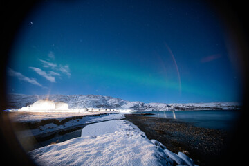 A dramatic night sky with Aurora Borealis over a row of illuminated glamping domes near a snowy coast and calm water