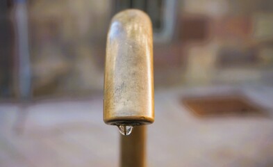 Brauner Wasserhahn mit Wassertropfen aus Kupfer über Brunnen af Platz in Stadt vor rotbraun-gelber Betonwand 