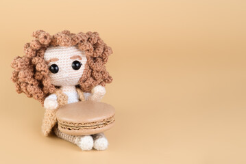 Handmade knitted doll with curly hair and macaron on beige surface, copy space.