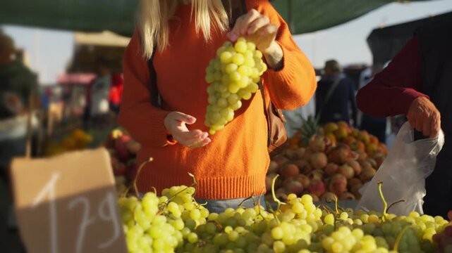 Blonde woman wear an orange sweater carefully select bunch of fresh green grapes from vendor's stall at bustling local farmers market on sunny day with warm, inviting light