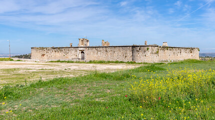 The medieval stone wall and entrance of the weathered Castillo de los Condes in Chinchón, Spain, framed by a lively field of yellow wildflowers and surrounded by a dirt road