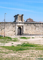 The medieval stone wall and entrance of the weathered Castillo de los Condes in Chinchón, Spain, framed by a lively field of yellow wildflowers and surrounded by a dirt road