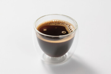 A transparent double-layer glass coffee cup filled with rich dark brown liquid, featuring a dense layer of fine foam on top with tiny bubbles clinging to the inner wall
