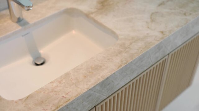 Close up of modern countertop with built in rectangular white sink. Surface is light colored possibly marble or granite with soft veining. Sleek faucet mounted at top left