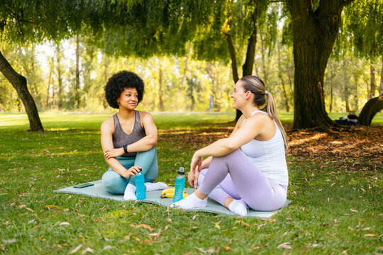 Two women relaxing on yoga mats, talking and smiling during their outdoor workout cool down - Powered by Adobe