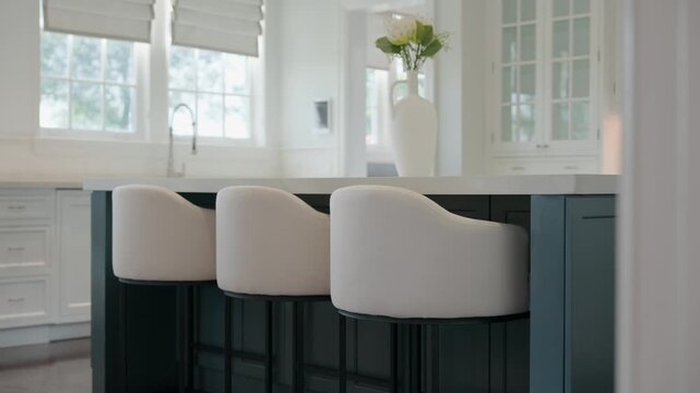 Stylish modern kitchen interior centers around sleek kitchen island with white countertop and dark cabinets. Three white upholstered bar stools with curved backs