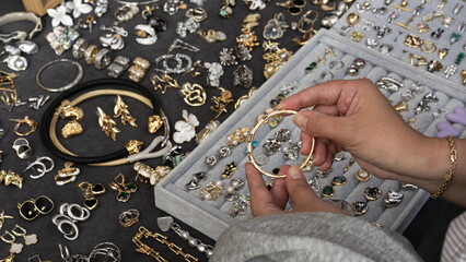Close-up of woman’s hands holding golden bracelet at jewelry stand, variety of silver and gold rings, earrings, and accessories on display
