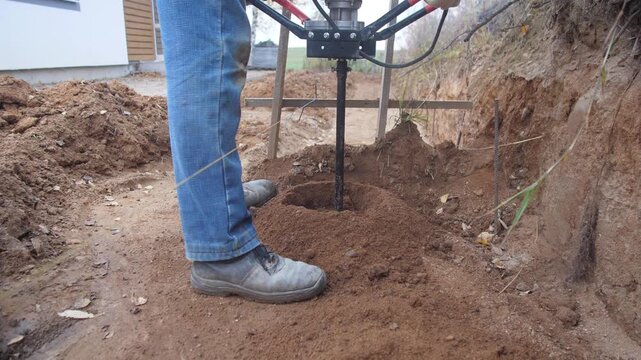 installer drilling deck post holes beside house, closeup of boots and jeans on packed soil, auger bit removed showing earth and footing, careful measurement and alignment for structure