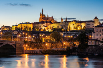 Prague Castle illuminated at blue hour with reflections on the Vltava River and city lights glowing in the evening sky.
