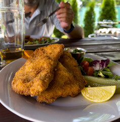 Crispy breaded schnitzel with lemon and mixed salad on white plate, man eating lunch at rustic outdoor restaurant table