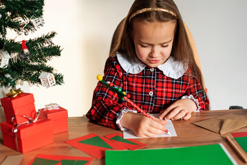 Young caucasian girl writing christmas cards by tree in plaid dress, Christmas wish letter to Santa...