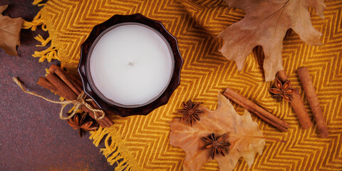 Top view of white candle in glass jar with cinnamon sticks, star anise and autumn leaves on yellow fabric