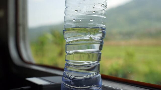 Close-up of a plastic water bottle on a train window with a scenic green landscape in the background.