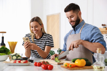 Cooking process. Smiling man cutting cucumber while his wife looking at recipe on smartphone at table in kitchen