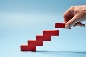 A white man's hand constructs a staircase with steps from wooden blocks. Conceptualization of growth, success, business progress, and goal achievement.