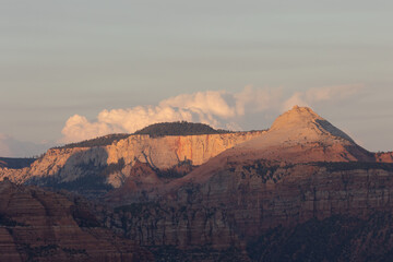 The mountains of Zion NP Utah USA seen from Smith Mesa road, glow in the warm light of sunset while puffy clouds drift past the rugged peaks and shadows creep up from the lower cliffs.