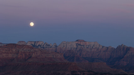 A full moon rises at sunset from behind the Altar of Sacrifice in Zion NP Utah USA, partly hidden by high thin clouds colored by the Belt of Venus.