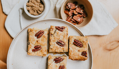 A plate of four pieces of dessert with a bowl of nuts in the background