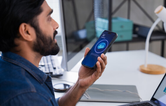Indian man using AI voice assistant on smartphone in office, interacting with virtual assistant app for productivity, AI-powered speech, and smart technology. - Powered by Adobe