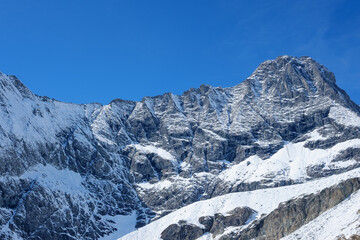 A stunning close-up view of majestic alpine peaks, newly blanketed in fresh, pristine snow from the first fall. Under a brilliant, cloudless sunny sky, the rugged beauty of the mountains is emphasized