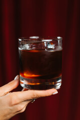 Dark and elegant cocktail in a glass with ice and a piece of chocolate, held by a hand on a dark red background. Sophisticated drink concept, stylish bar or lounge atmosphere.