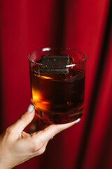 Dark and elegant cocktail in a glass with ice and a piece of chocolate, held by a hand on a dark red background. Sophisticated drink concept, stylish bar or lounge atmosphere.