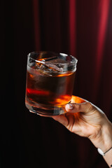 Dark and elegant cocktail in a glass with ice and a piece of chocolate, held by a hand on a dark red background. Sophisticated drink concept, stylish bar or lounge atmosphere.