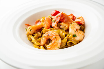 A delicious bowl of pappardelle pasta with fresh shrimp, halved cherry tomatoes, and a savory sauce, garnished with herbs. The dish is presented on a white plate with a neutral background.