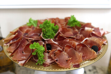 Thinly sliced prosciutto served on a golden plate with lettuce leaves and black olives for a festive table.
