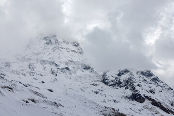 The snow-covered peaks of the Himalayas are partially obscured by thick clouds. The landscape of Annapurna Sanctuary is visible in Nepal.