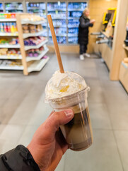 Person's hand holding a to-go iced coffee with whipped cream and a paper straw in a convenience store.