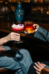 Two people clinking glasses with cocktails at a bar. Red floral drink and dark whiskey cocktail in elegant glasses, celebrating in a cozy evening atmosphere.
