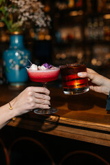 Two people clinking glasses with cocktails at a bar. Red floral drink and dark whiskey cocktail in elegant glasses, celebrating in a cozy evening atmosphere.
