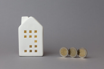 White ceramic house model beside three coins on a gray background, symbolizing finance, investment, and home ownership concepts. copy space