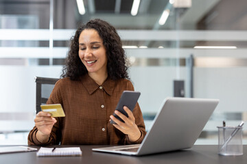 Smiling young woman holding credit card and smartphone while completing an online purchase at a desk with laptop and tablet, remote work setup and secure mobile payment visible