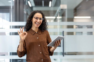 Businesswoman with curly hair and glasses holding a digital tablet, smiling and giving an optimistic okay gesture, symbolizing success and approval in a modern office environment